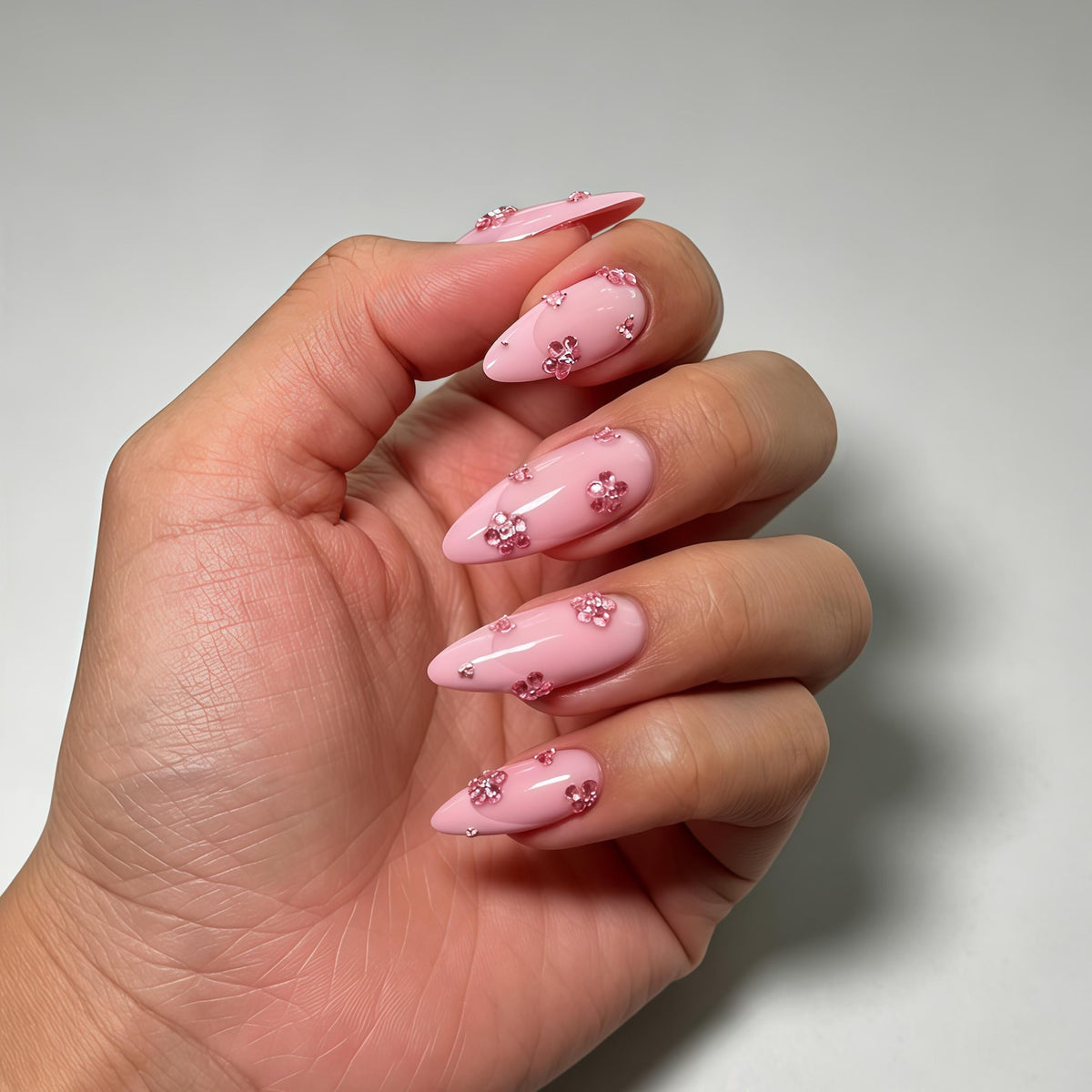 Almond-shaped nails with a glossy light pink base coat. Each nail is decorated with small, sparkling pink floral-shaped rhinestones arranged in clusters across the surface, creating a textured and reflective appearance.