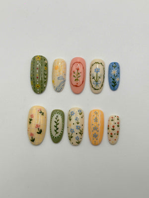 A set of ten oval-shaped press-on nails featuring various floral and botanical designs. The nails display a palette of green, cream, pink, blue, and orange, with intricate hand-painted patterns including stripes, grids, and frames.