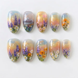 A set of ten almond-shaped press-on nails featuring a hand-painted landscape design. The artwork displays a soft sunset sky background with various floral motifs including purple lupines, white daisies, and orange wildflowers.