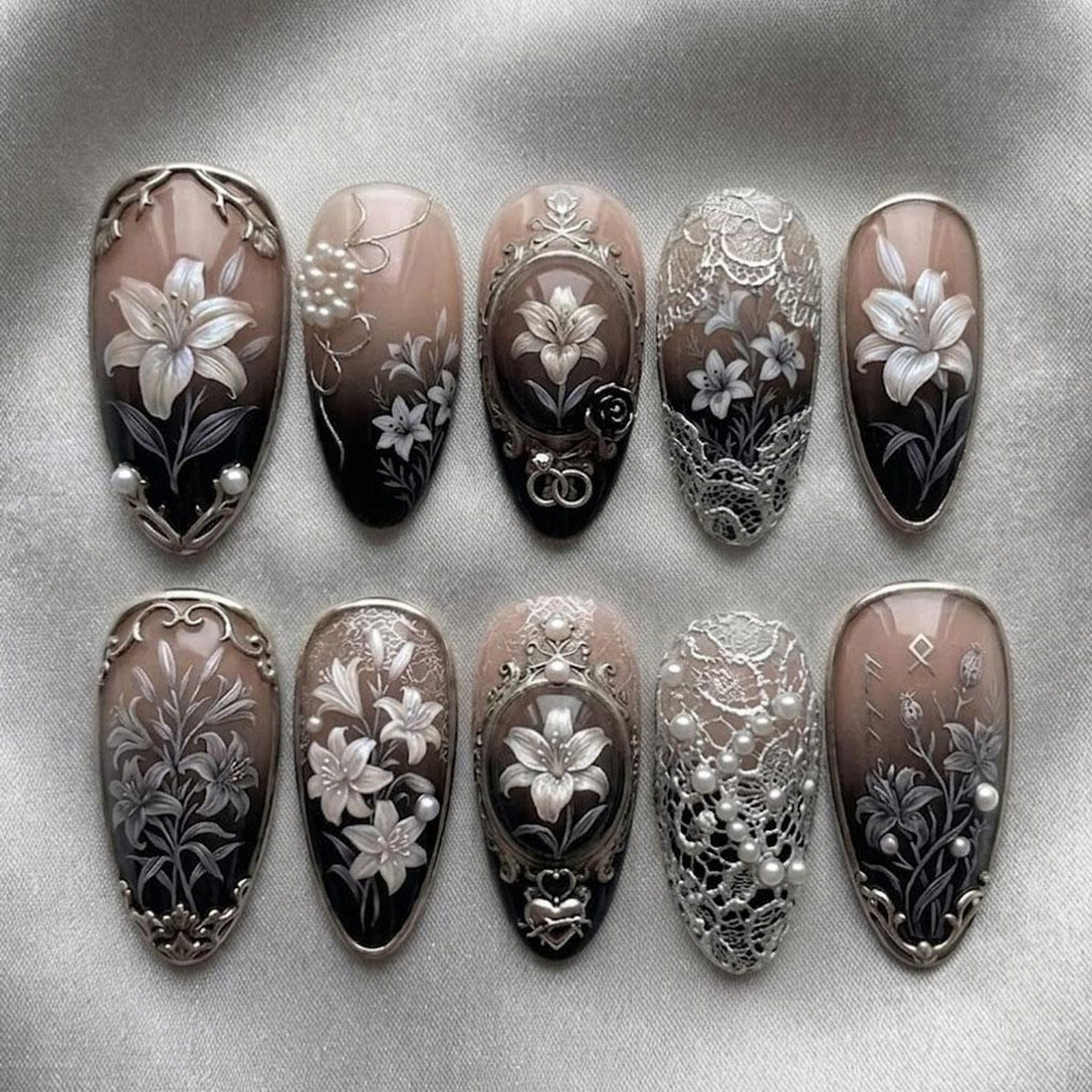 A set of ten press-on nails featuring a gradient from nude to black. The designs include white lily illustrations, silver filigree accents, raised pearl embellishments, and intricate lace patterns, all finished with a metallic silver trim.