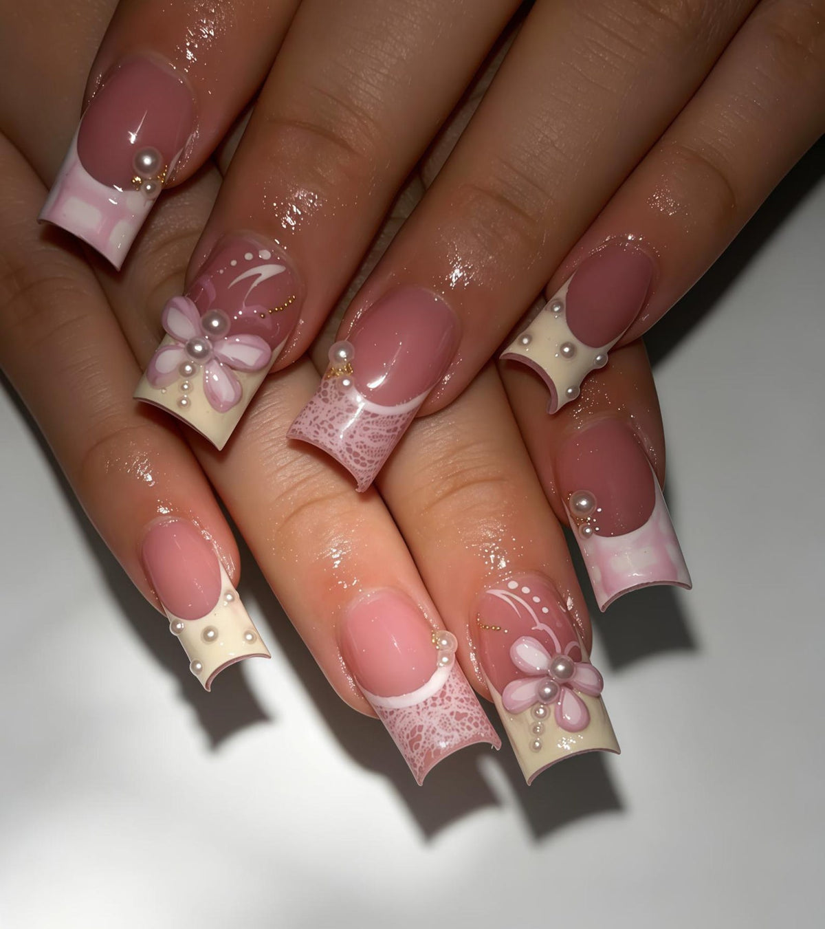 Square-shaped artificial nails featuring a French manicure base with a nude pink nail bed. The tips are decorated with white lace patterns, raised pink flower petals, small white pearls, and thin gold lines on a cream-colored background.