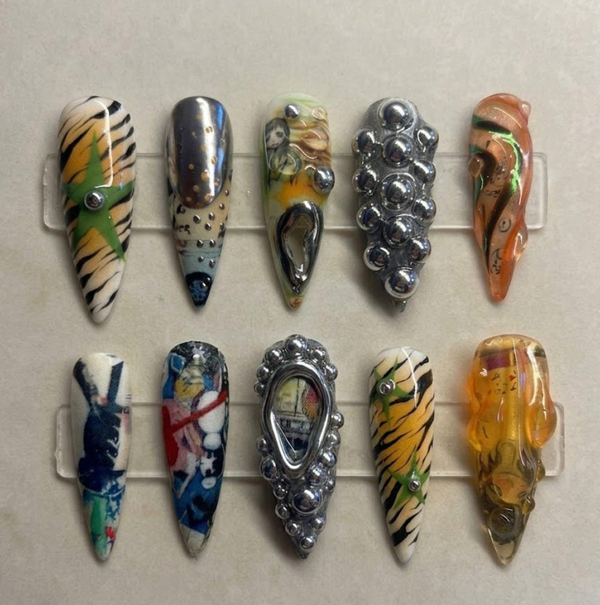 A set of ten almond-shaped press-on nails featuring diverse designs including tiger stripes with green stars, metallic silver studs, abstract multi-colored patterns, 3D silver spheres, and translucent amber resin with embedded details.