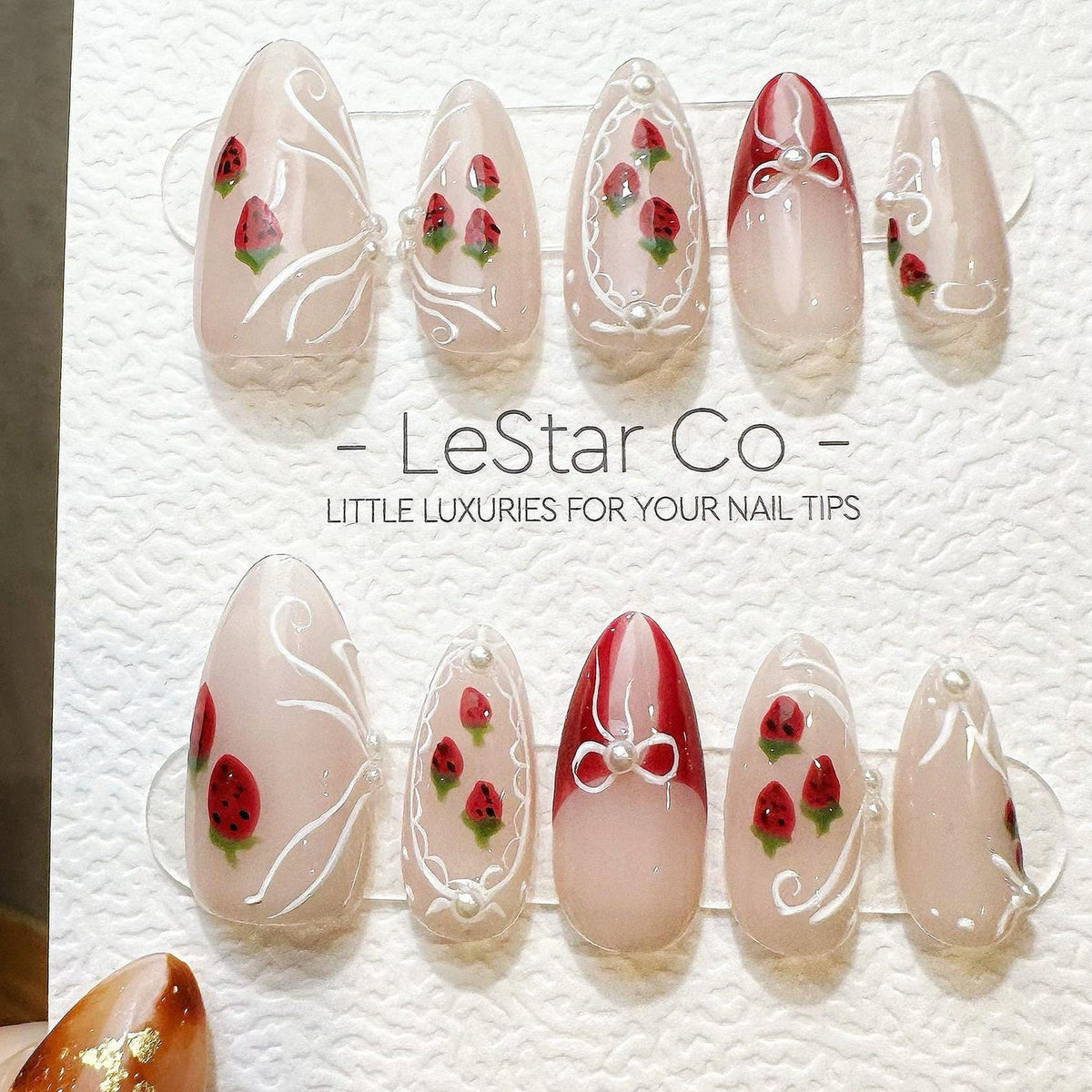 A set of ten almond-shaped press-on nails featuring a sheer nude base with red strawberry illustrations, white swirl accents, and small white pearl embellishments. The display card includes the text L