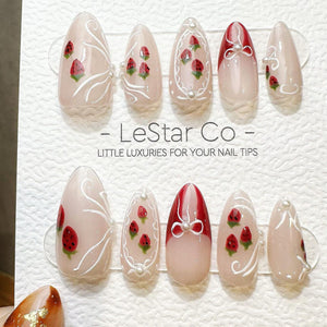 A set of ten almond-shaped press-on nails featuring a sheer nude base with red strawberry illustrations, white swirl accents, and small white pearl embellishments. The display card includes the text L