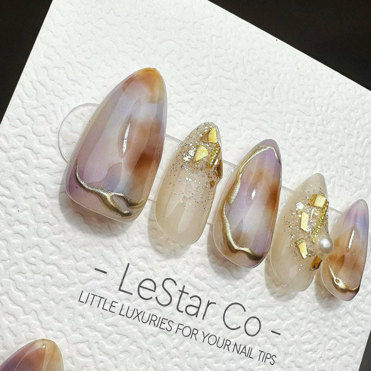 Five almond-shaped press-on nails featuring a marbled design in shades of lavender, cream, and brown with gold foil accents and a single pearl. The nails are displayed on a white card printed with the