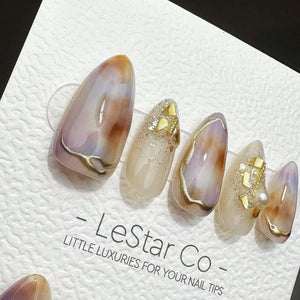 Five almond-shaped press-on nails featuring a marbled design in shades of lavender, cream, and brown with gold foil accents and a single pearl. The nails are displayed on a white card printed with the
