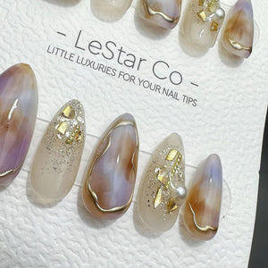 A set of press-on nails featuring a marbled design in shades of beige, brown, and soft purple, accented with gold foil flakes, silver glitter, and small white pearls. The display card reads LeStar Co 