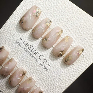 A set of ten press-on nails with a translucent nude base, featuring gold foil flakes, pink circular accents, and thin white marbled lines. The nails are displayed on a white textured card labeled LeSt