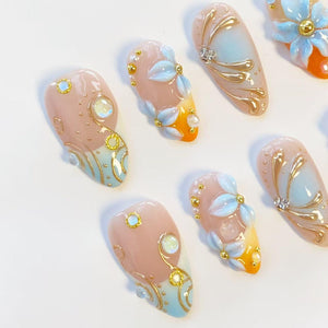 A set of almond-shaped press-on nails featuring a gradient of nude, soft blue, and orange. The designs include raised 3D light blue flower petals, metallic gold swirling lines, small gold beads, and iridescent circular rhinestones.