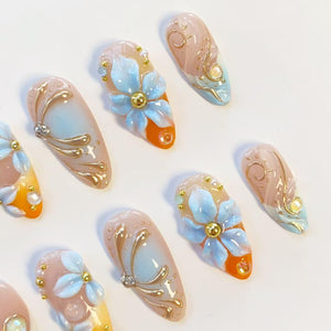 A set of almond-shaped press-on nails featuring a gradient of peach, orange, and light blue. The designs include three-dimensional light blue floral petals, gold metallic swirls, small gold beads, and clear rhinestones.