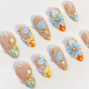 A set of ten almond-shaped press-on nails featuring a gradient of nude, light blue, and orange. The designs include raised 3D light blue flower petals, metallic gold swirling accents, small gold beads, and iridescent rhinestones.