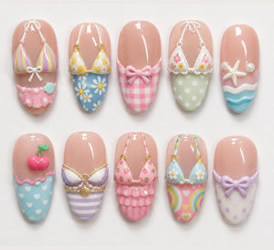 A set of ten press-on nails featuring 3D designs including bikinis with floral, striped, and gingham patterns, a starfish, cherries, and bows. The nails have a glossy finish with various pastel colors like pink, blue, and yellow.