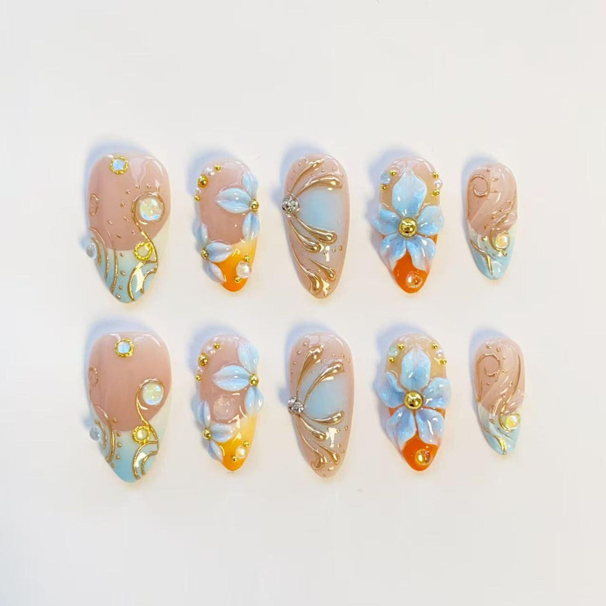 A set of ten almond-shaped press-on nails featuring a nude base with gradient orange and blue tips. The designs include raised 3D light blue flowers, gold metallic swirls, small white pearls, and iridescent rhinestones on each nail.