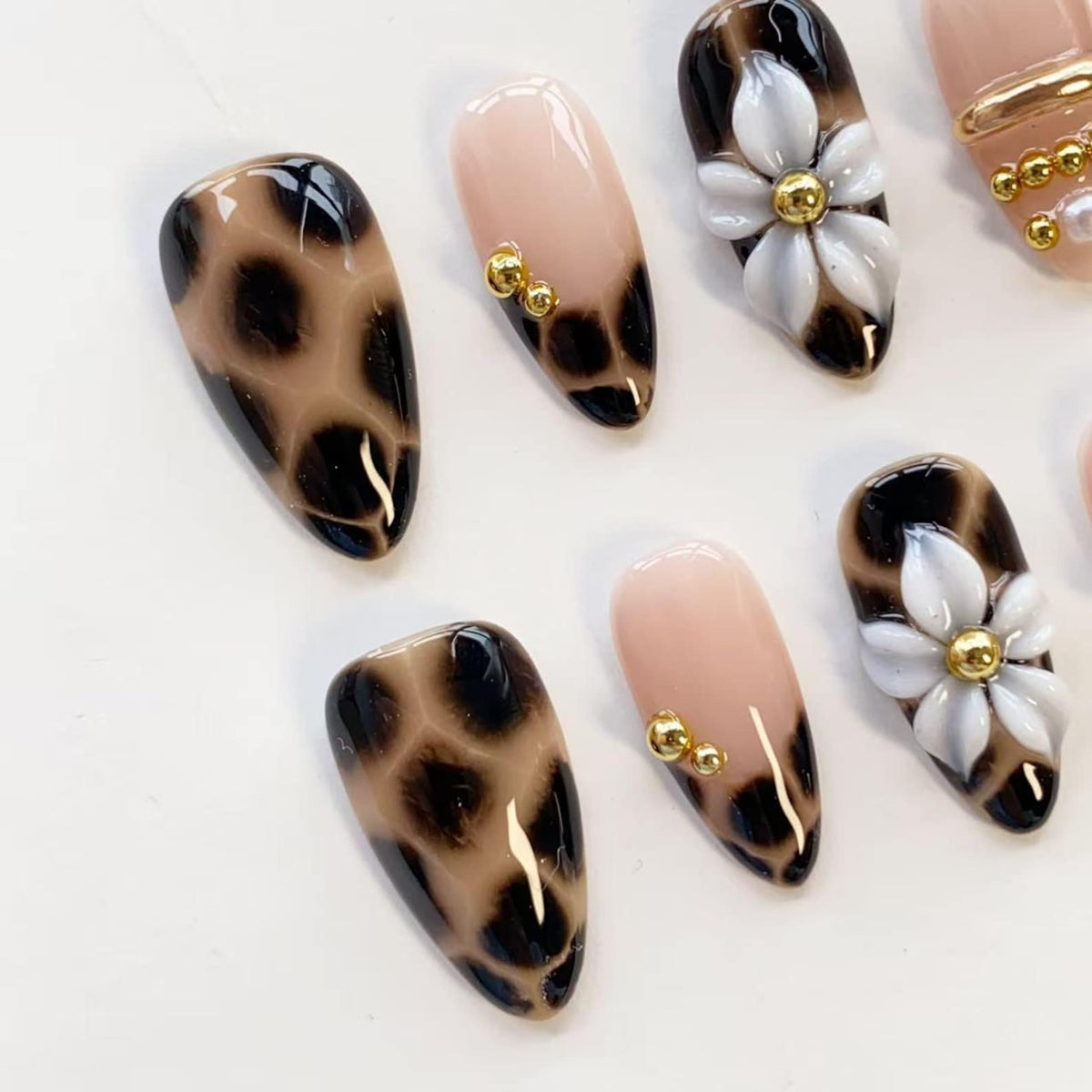 A set of almond-shaped press-on nails featuring a mix of designs including a brown and black leopard print, solid nude bases with small gold metallic spheres, and nude bases adorned with white 3D floral accents and gold centers.