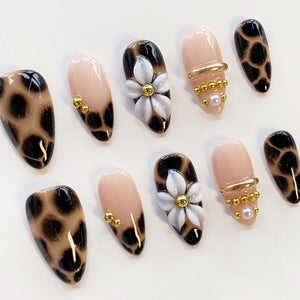 A set of ten press-on nails featuring a mix of designs including a brown and black leopard print, solid nude base with gold beads, and nude nails adorned with a white floral accent, gold metallic bars, and small white pearls.