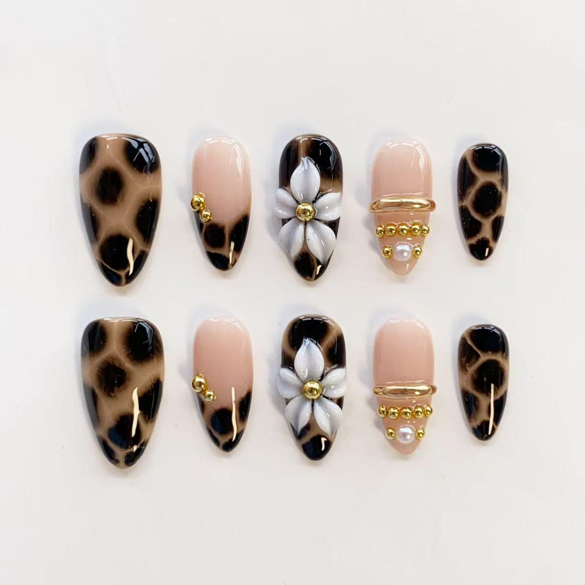 A set of ten press-on nails featuring a mix of designs. Some nails have a brown and black leopard print, while others are a solid nude color accented with white 3D flowers, small gold metallic beads, and tiny white pearls.