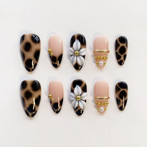 A set of ten press-on nails featuring a mix of designs. Some nails have a brown and black leopard print, while others are a solid nude color accented with white 3D flowers, small gold metallic beads, and tiny white pearls.