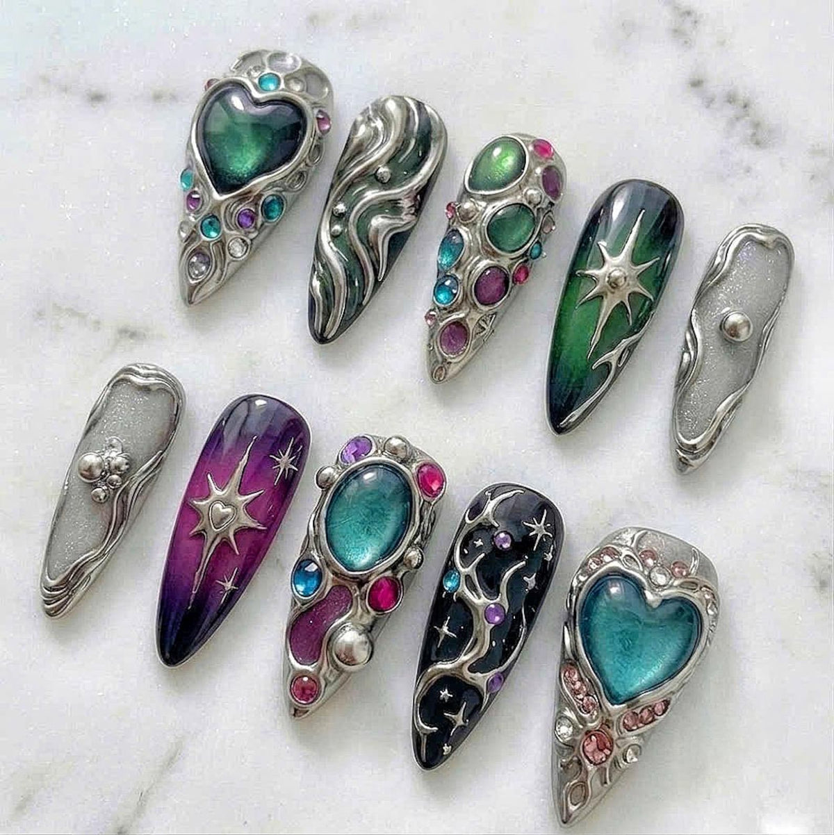 A set of ten press-on nails featuring metallic silver accents, sculpted liquid metal textures, and embedded colorful gemstones. The designs include heart shapes, starburst motifs, and gradient color transitions in shades of green and purple.