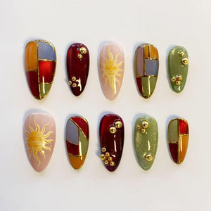 A set of ten almond-shaped press-on nails featuring a variety of designs including color-blocked segments in red, orange, olive green, and grey with gold lines, sun motifs on light pink bases, and deep red or green with gold beads.