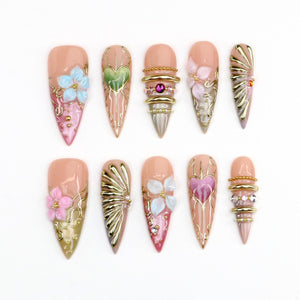 A set of ten almond-shaped press-on nails featuring a nude base with intricate 3D designs. Decorations include raised pink and blue flowers, metallic gold filigree, heart motifs, gold rings, and small embedded crystals.