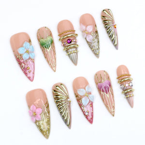 A set of ten stiletto-shaped press-on nails featuring a nude base with intricate 3D designs including gold metallic accents, raised floral motifs in pink and blue, heart shapes, and embedded rhinestones in various sizes and colors.