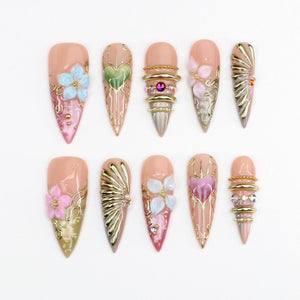 A set of ten stiletto-shaped press-on nails featuring a nude base with intricate 3D embellishments including gold metallic lines, raised floral accents in blue and pink, heart motifs, and small embedded crystals and gold beads.