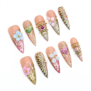 A set of ten stiletto-shaped press-on nails with a nude base, featuring intricate 3D gold metallic accents, raised floral designs in pink, blue, and white, heart-shaped motifs, and embedded rhinestones in various colors and sizes.