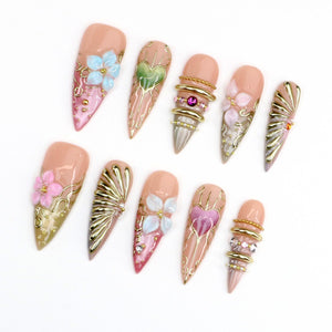 A set of ten stiletto-shaped press-on nails featuring a nude base with intricate 3D embellishments, including light blue and pink floral accents, metallic gold filigree, heart motifs, and small embedded crystals and gold beads.