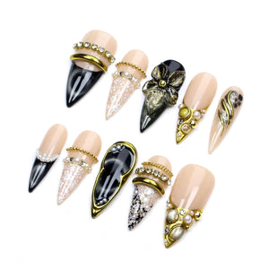 A set of ten stiletto-shaped press-on nails featuring a nude base with various decorative elements including black cat-eye polish, white lace patterns, gold metallic accents, clear rhinestones, small white pearls, and a three-dimensional black flower