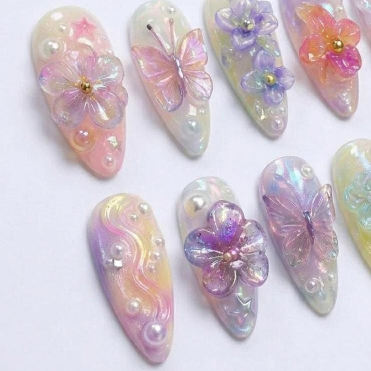 A set of almond-shaped press-on nails featuring iridescent pastel gradients, 3D translucent butterfly and flower charms, small white pearls, and tiny star-shaped accents. Each nail displays a glossy finish with layered textures.