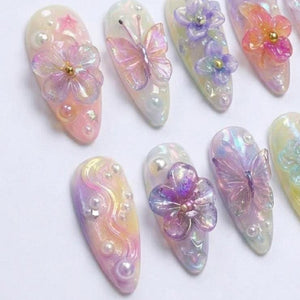 A set of almond-shaped press-on nails featuring iridescent pastel gradients, 3D translucent butterfly and flower charms, small white pearls, and tiny star-shaped accents. Each nail displays a glossy finish with layered textures.