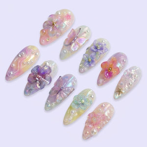 A set of ten almond-shaped press-on nails featuring iridescent pastel finishes, adorned with three-dimensional acrylic flowers, butterflies, small white pearls, and tiny gold star charms arranged in various decorative patterns.