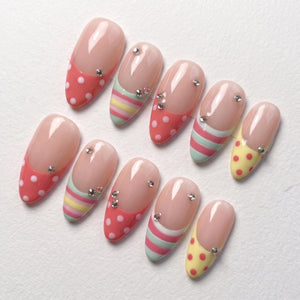 A set of ten press-on nails featuring a nude base with colorful tips. The designs include coral pink with white polka dots, pastel striped patterns in mint, yellow, and pink, and solid yellow with red dots, all accented with small clear rhinestones.