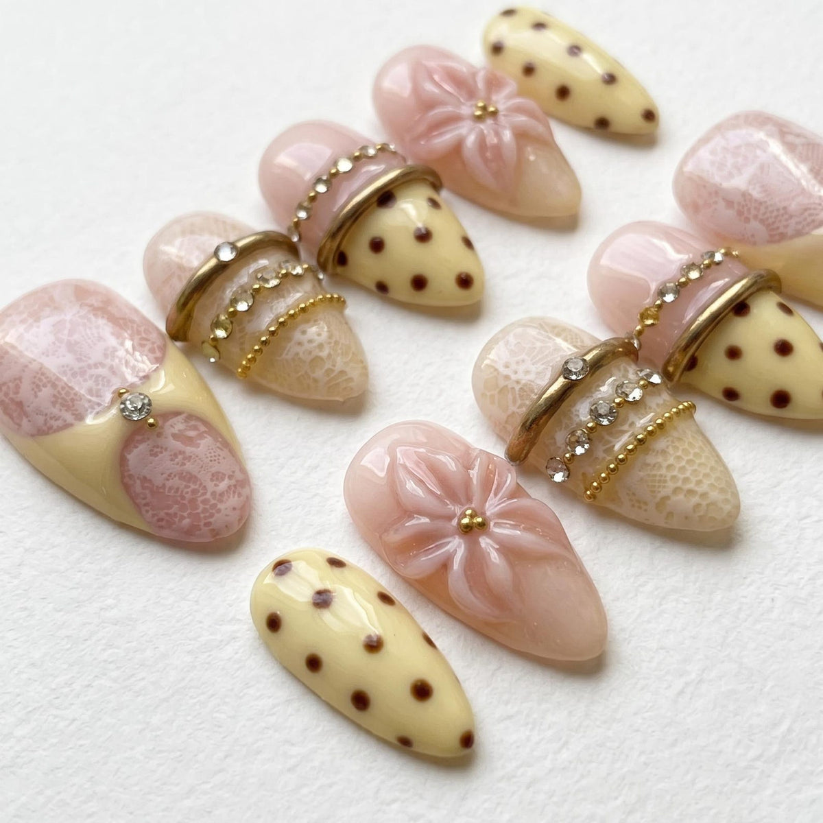 A set of almond-shaped press-on nails featuring a mix of designs including cream-colored nails with brown polka dots, pale pink nails with white lace patterns, and nails adorned with 3D pink floral accents, gold bands, and rhinestones.