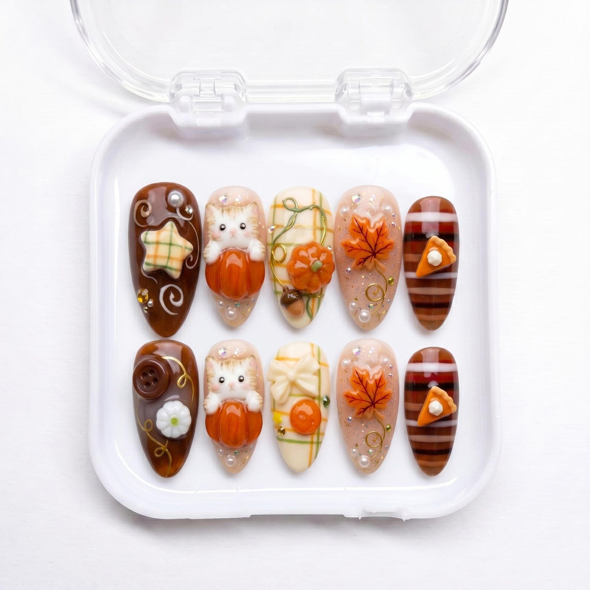 A set of ten almond-shaped press-on nails in a white plastic case. The designs feature autumn themes including 3D orange pumpkins, kittens, maple leaves, plaid patterns, brown buttons, and small slices of pumpkin pie with white cream.