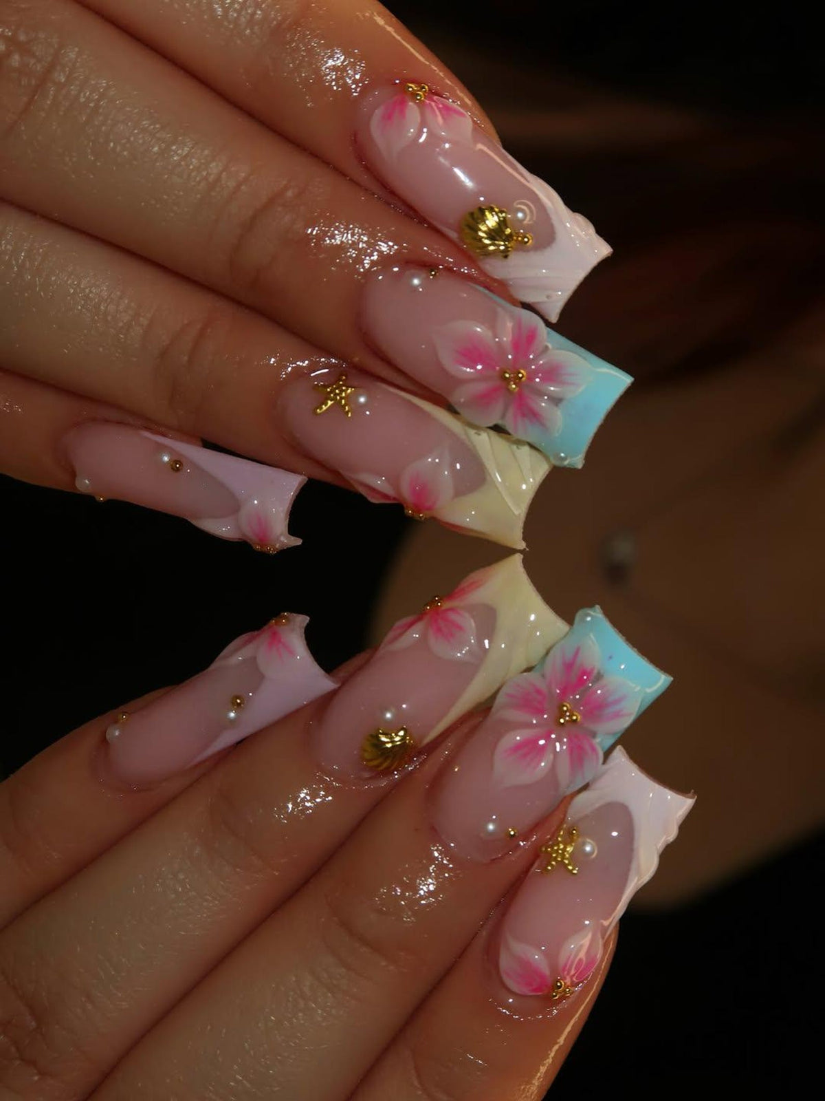 Long square-shaped artificial nails featuring a nude base with pink floral hand-painted designs. The nail art includes three-dimensional gold seashell and starfish charms, along with small white pearl accents and light blue tips.
