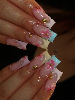 Long square-shaped artificial nails featuring a nude base with pink floral hand-painted designs. The nail art includes three-dimensional gold seashell and starfish charms, along with small white pearl accents and light blue tips.