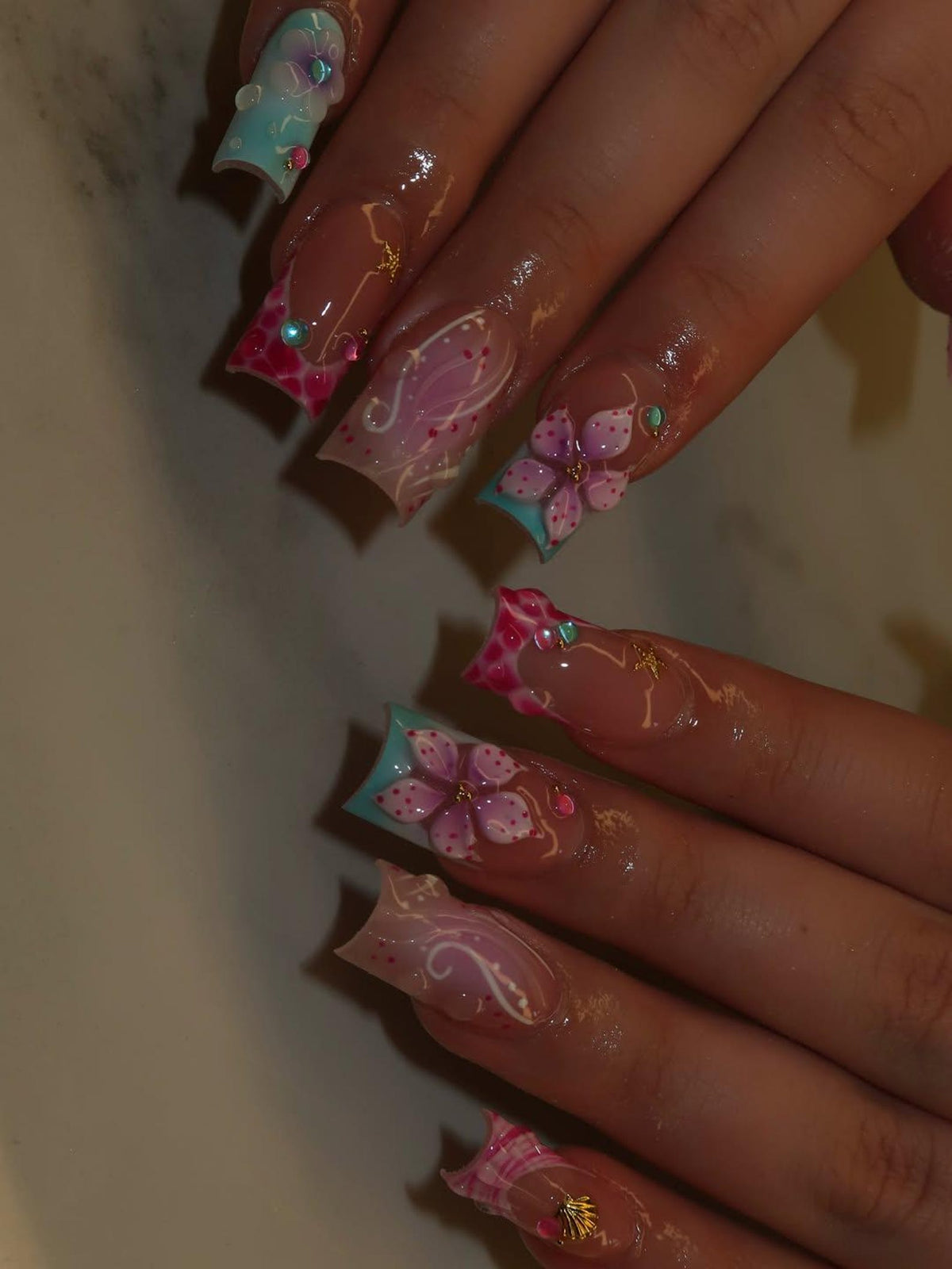Long square-shaped artificial nails featuring a mix of light blue and pink tips. The designs include hand-painted purple flowers, white swirling line art, small gold starfish charms, and tiny colorful beads embedded on the nail surface.