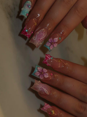 Long square-shaped artificial nails featuring a mix of light blue and pink tips. The designs include hand-painted purple flowers, white swirling line art, small gold starfish charms, and tiny colorful beads embedded on the nail surface.
