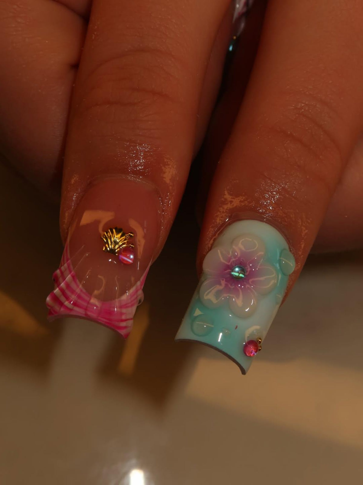Two manicured fingernails featuring square tips. One nail has a clear base with a pink plaid pattern at the tip and a small gold seashell charm. The other nail features a light blue gradient base with a raised white flower and pink gems.