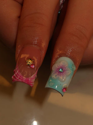 Two manicured fingernails featuring square tips. One nail has a clear base with a pink plaid pattern at the tip and a small gold seashell charm. The other nail features a light blue gradient base with a raised white flower and pink gems.