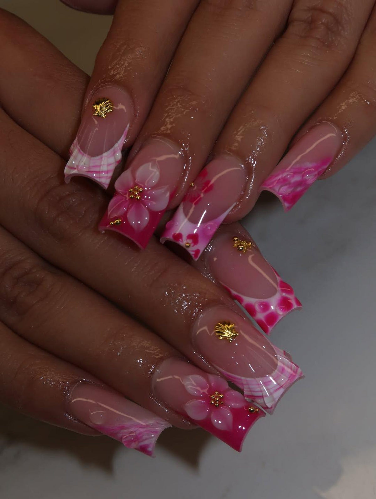 Long square-shaped artificial nails featuring a French manicure style with a translucent base. The tips are decorated with vibrant pink floral patterns, white accents, and small gold-colored metallic shell and bead embellishments.