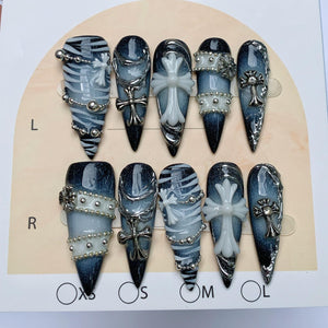 A set of ten stiletto-shaped press-on nails featuring a dark blue and black gradient base. The designs include white zebra-like stripes, silver metallic chains, small silver beads, and raised white or silver cross emblems.