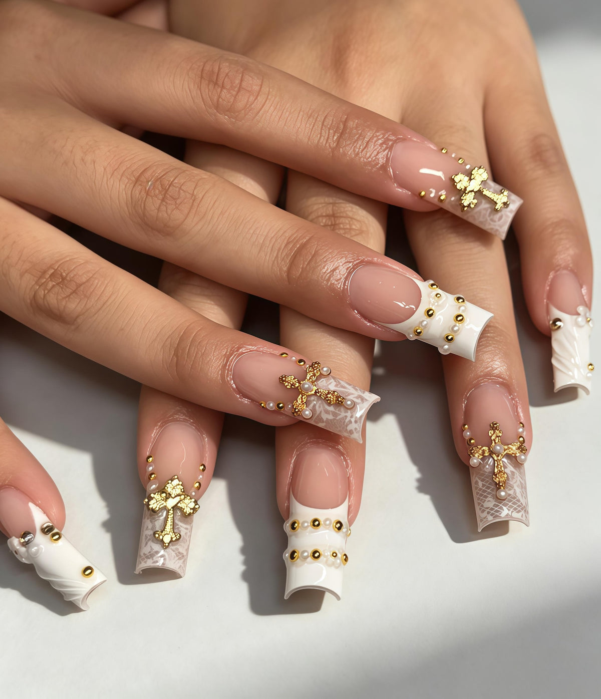 Long square-shaped artificial nails featuring a French manicure base with white lace patterns. The nails are decorated with gold cross charms, small white pearls, and tiny gold metallic beads arranged in various patterns across the tips.