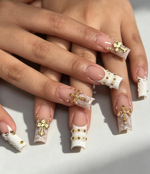 Long square-shaped artificial nails featuring a French manicure base with white lace patterns. The nails are decorated with gold cross charms, small white pearls, and tiny gold metallic beads arranged in various patterns across the tips.