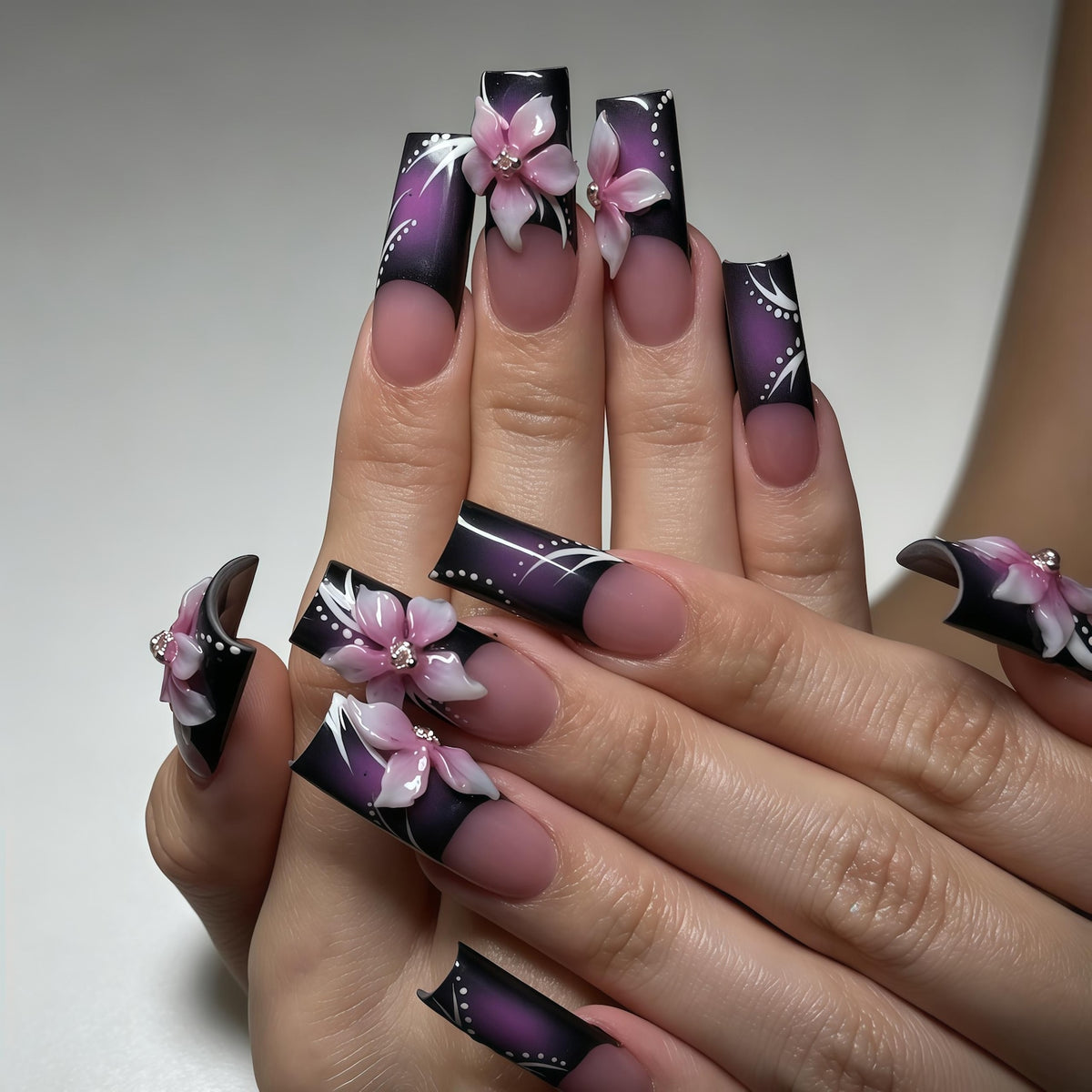 Long square-shaped artificial nails featuring a dark purple to black gradient base with a nude nail bed. The tips are decorated with three-dimensional light pink floral accents, small silver rhinestones, and delicate white line art.