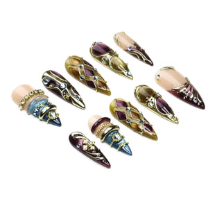 A set of ten stiletto-shaped press-on nails featuring a variety of designs including deep purple and gold lattice patterns, metallic accents, embedded rhinestones, and smooth nude-toned bases with ornate gold-toned embellishments.