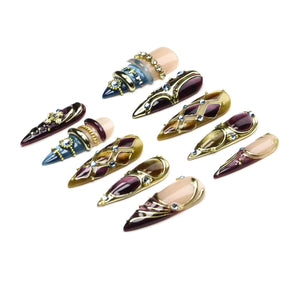 A set of ten stiletto-shaped press-on nails featuring a variety of designs, including deep purple, blue, and tan tones. The nails are decorated with gold-colored metal accents, intricate lattice patterns, and small embedded clear crystals.