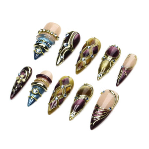 A set of ten press-on nails featuring pointed tips, decorated with gold-tone metal accents, embedded rhinestones, and a color palette of nude, deep purple, and blue. The designs include geometric patterns, layered rings, and metallic swirls.