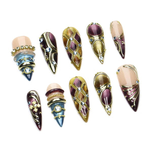 A set of ten press-on nails featuring almond and stiletto shapes. The designs include deep purple, gold, and blue tones with metallic accents, embedded rhinestones, cross charms, and intricate geometric patterns across the surfaces.